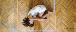 A young beautiful black-haired girl dressed in a white T-shirt and white shorts lying on her side in a room on a wooden parquet in a square painted with a white paint, bending her legs in a lap and pressing them against her chest and hugging them with a hands. Top view - How To Protect Yourself When Self-Isolating With A Controlling Partner - Sandra Harewood Counselling