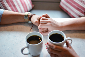 Couple Holding Hands Across Table With Cups Of Coffee - Sandra Harewood