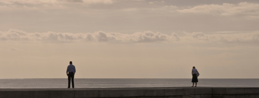 Couple standing apart looking out to sea, one partner wants to stay while the other feels unsure about the relationship